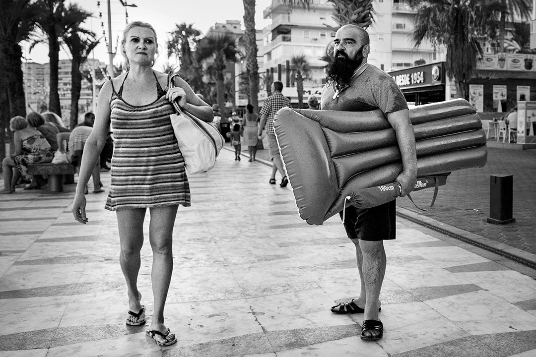 A woman in a striped dress and a man carrying a beach mattress on the promenade.